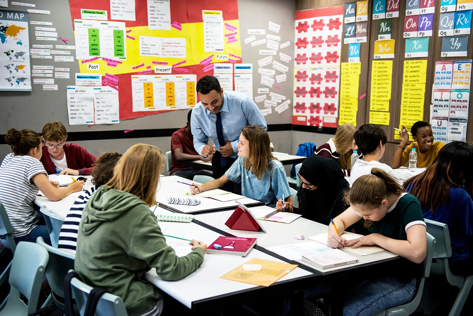 A classroom scene showing a diverse group of students engaged in learning. The teacher, a man in a light blue shirt and tie, is leaning over to assist a student.