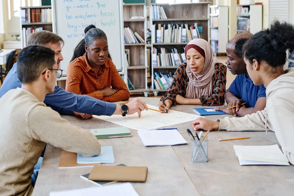 A diverse group of adults are seated around a table in what appears to be a library or classroom.