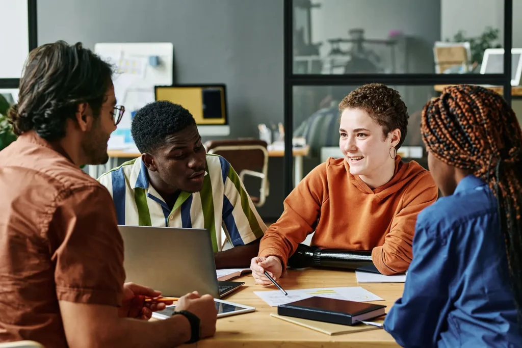 A diverse group of four young professionals or students are seated around a table in an office or workspace environment. They appear to be having a collaborative discussion or meeting.