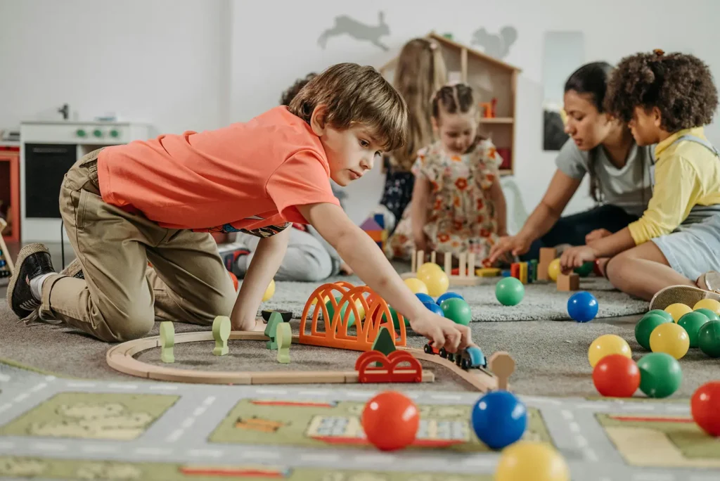 A group of young children are playing in a colorful preschool or daycare setting. In the foreground, a boy in an orange shirt and tan pants is kneeling on the floor, focused on playing with a wooden train set and bridge.